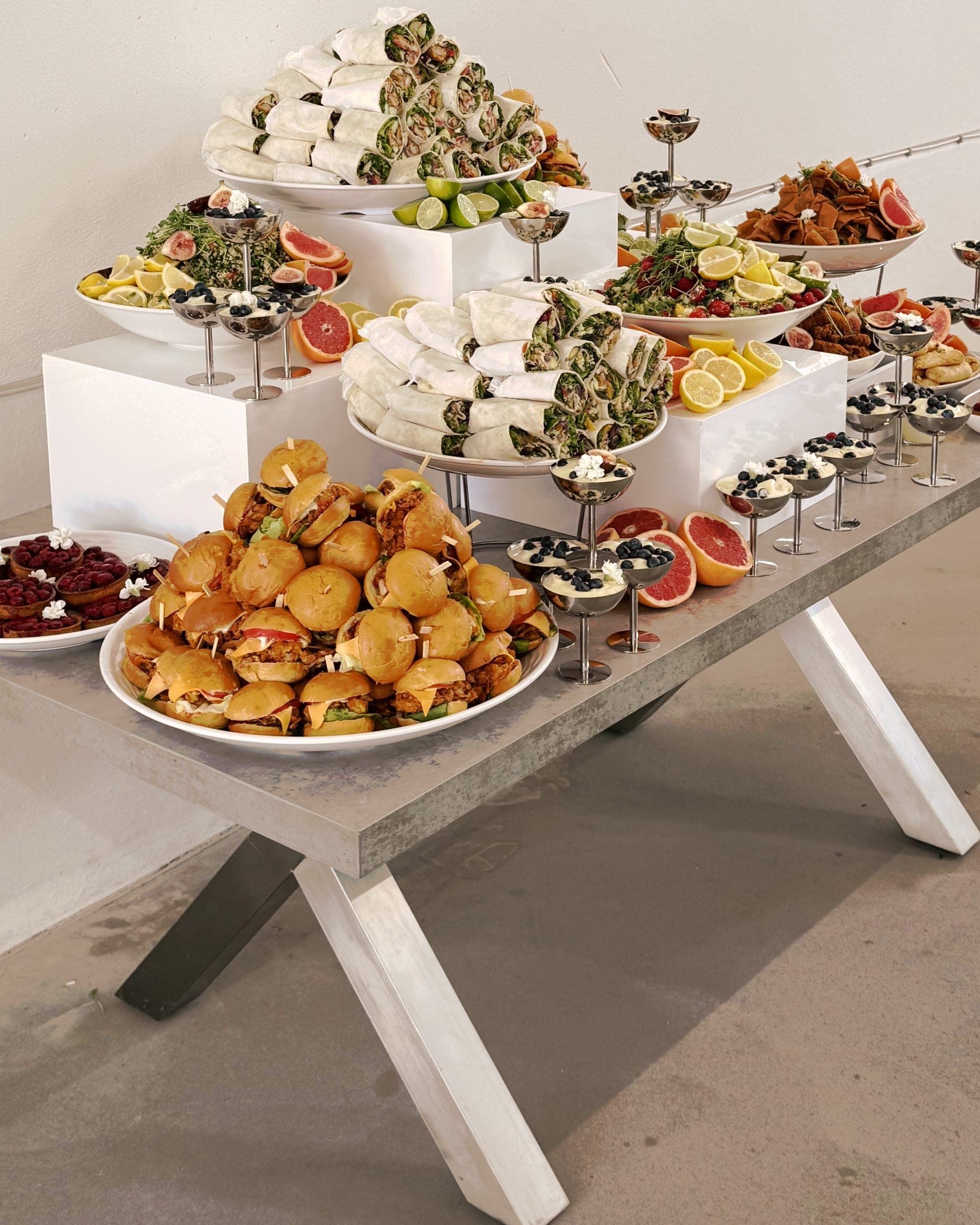Langer Buffettisch mit gekreuzten Stahlbeinen: gestapelte Wraps, ein Berg Mini-Burger, Salate mit Zitrusfrüchten und kleine Dessertschalen